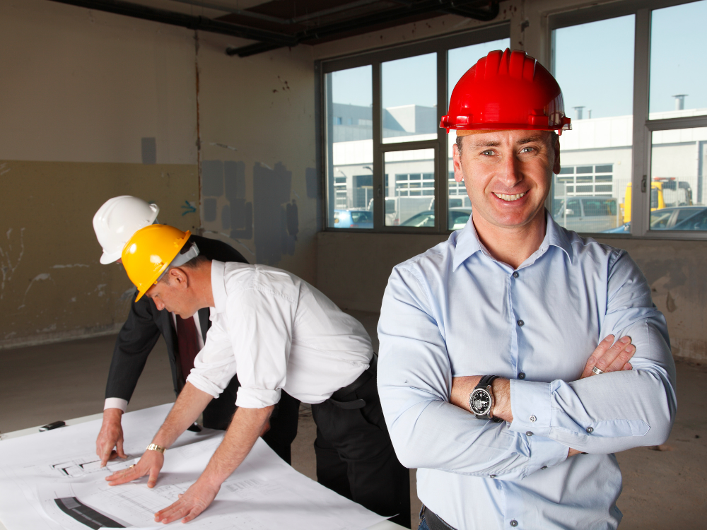 Man standing with arms crossed in a hard hat while two other men read over blueprint plans